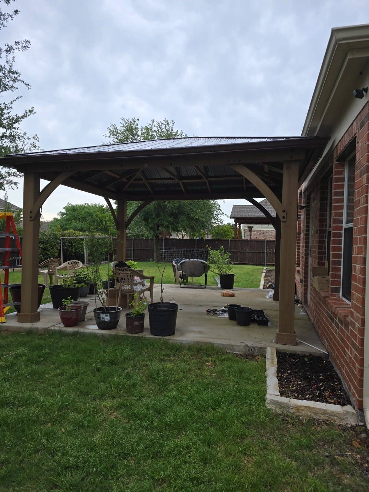 Wooden gazebo kit with a metal roof over a concrete patio beside a brick house
