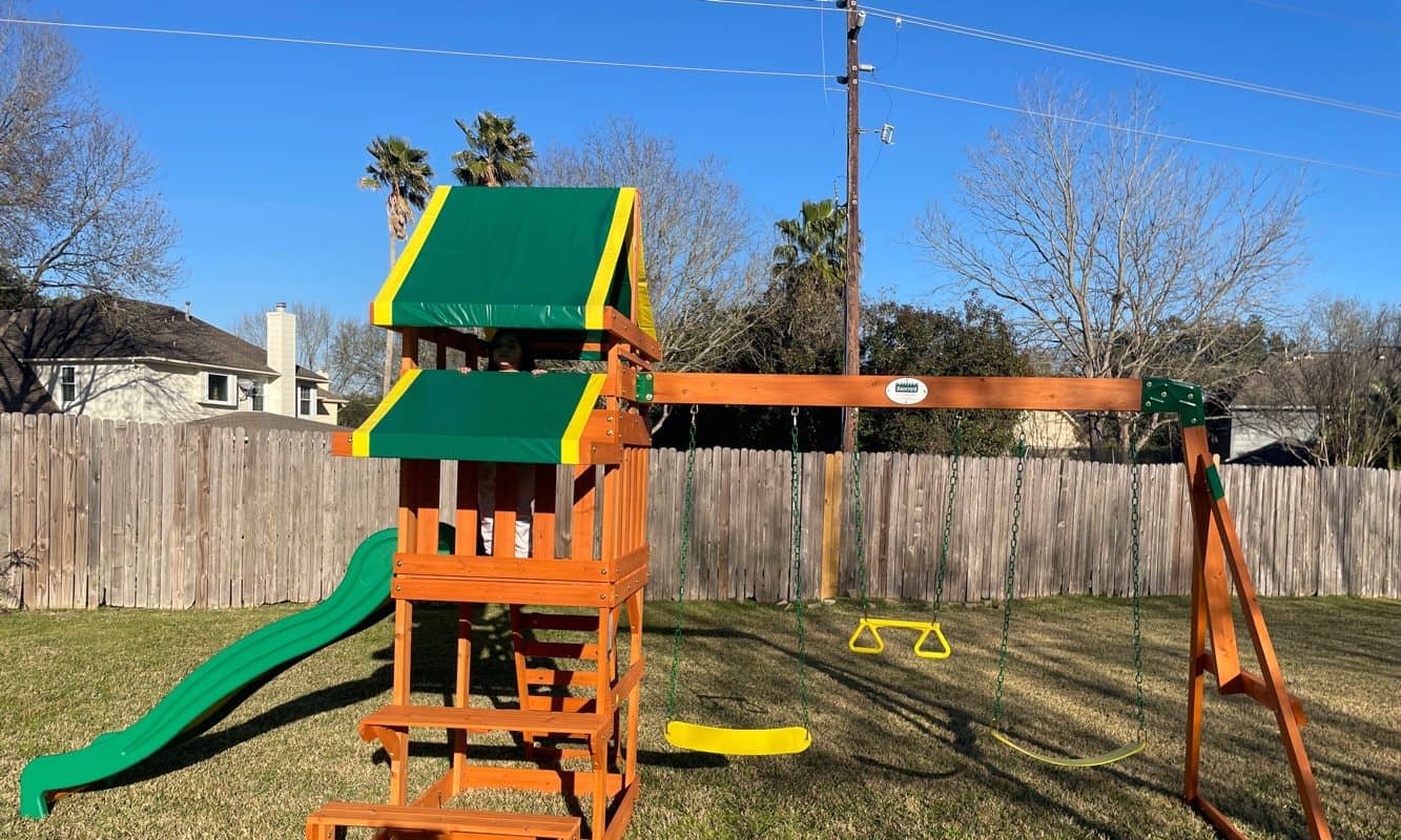 Wooden playset with a green slide, canopy and swing beam assembled in a fenced backyard