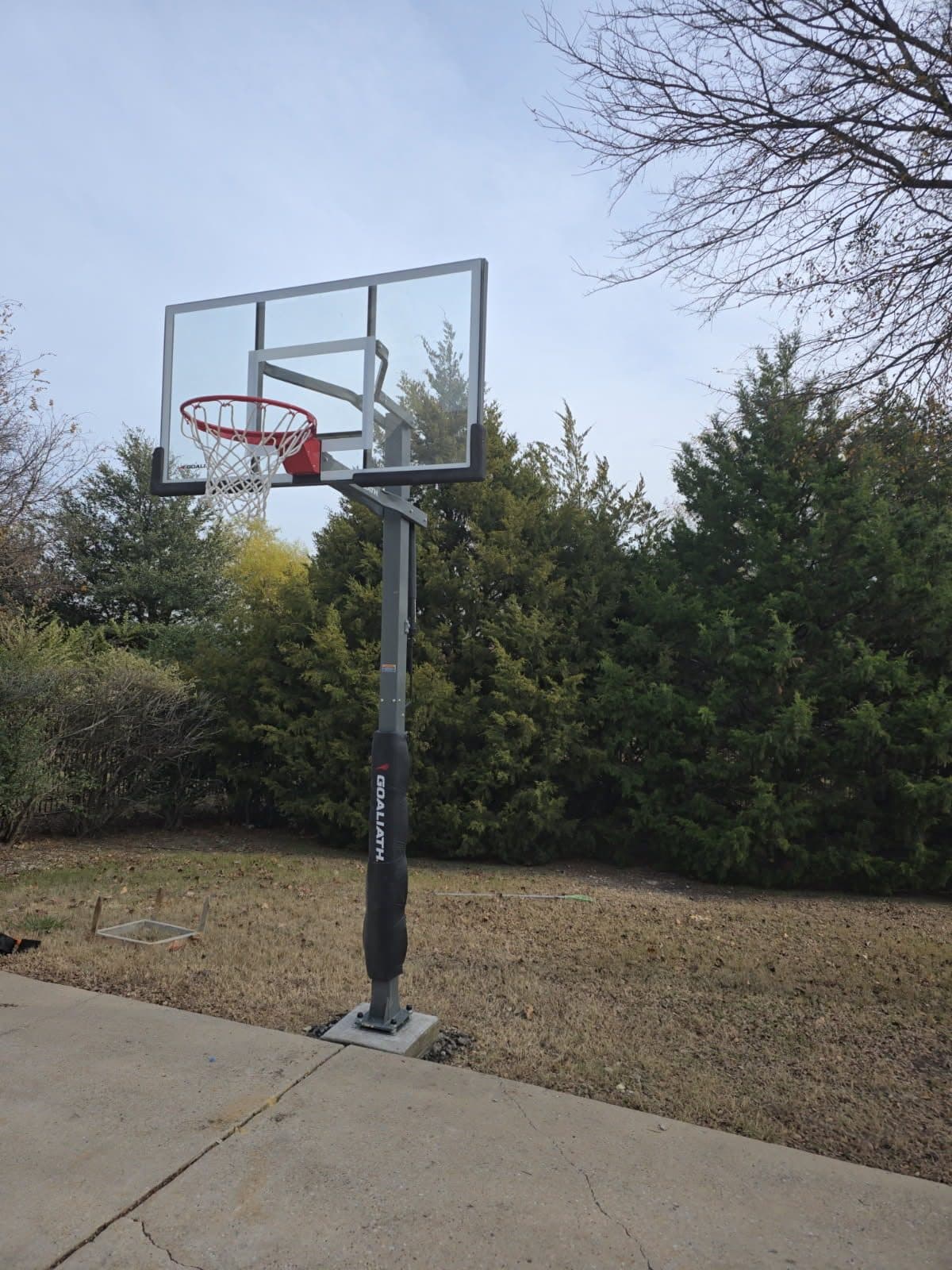 In-ground basketball goal with a clear backboard and padded pole, bolted to a concrete footing beside a driveway
