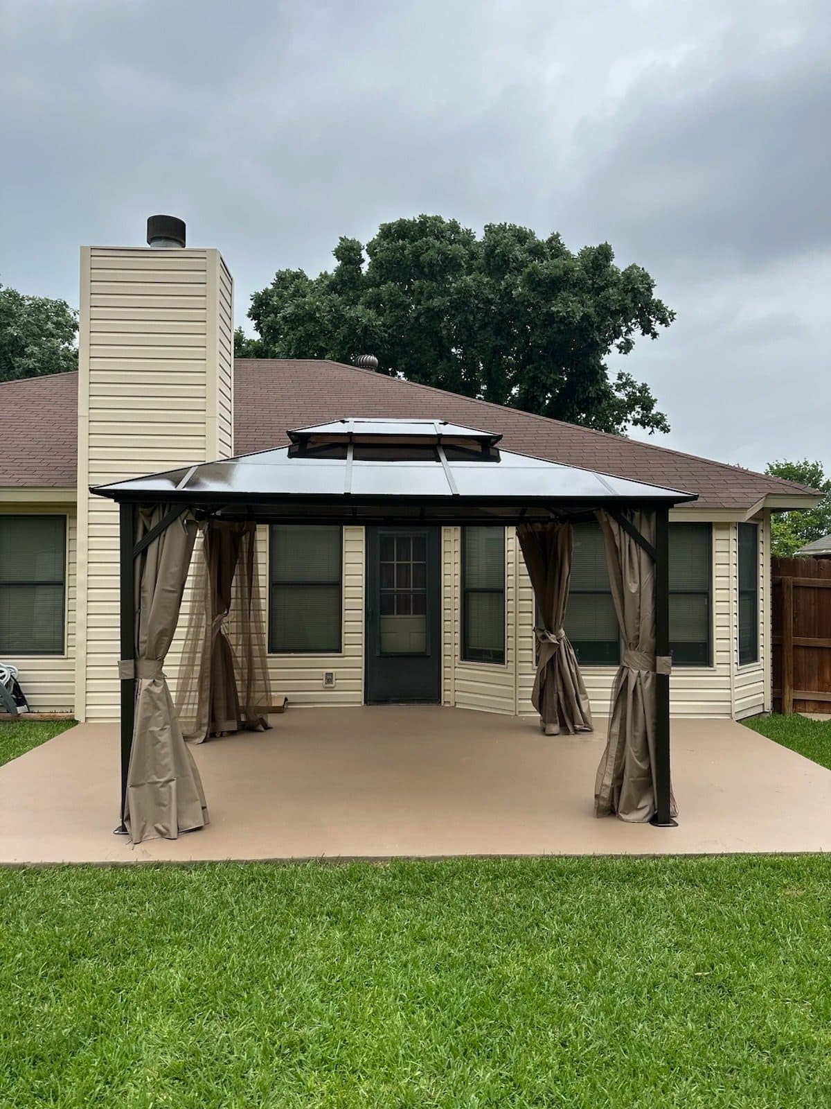 Double-roof metal gazebo with tan curtains tied back at each post on a backyard patio