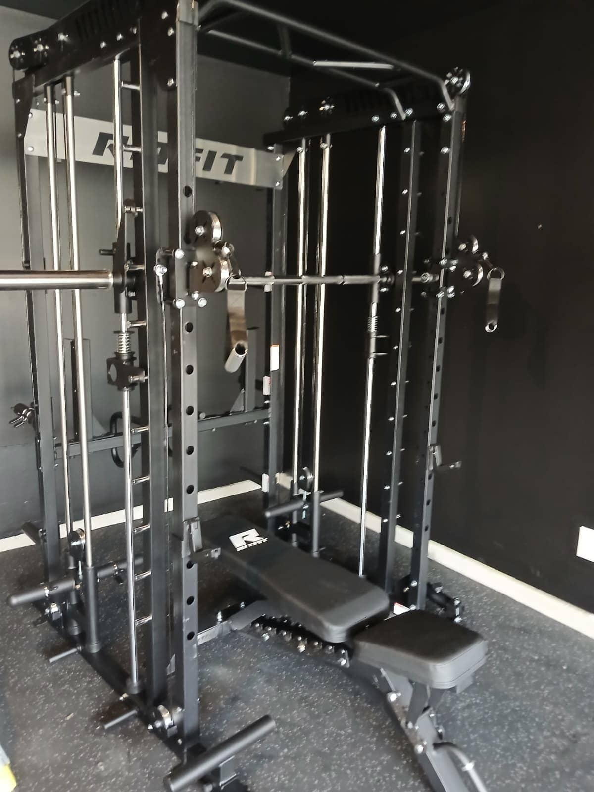 Smith machine and cable functional trainer with a bench, assembled against a black wall