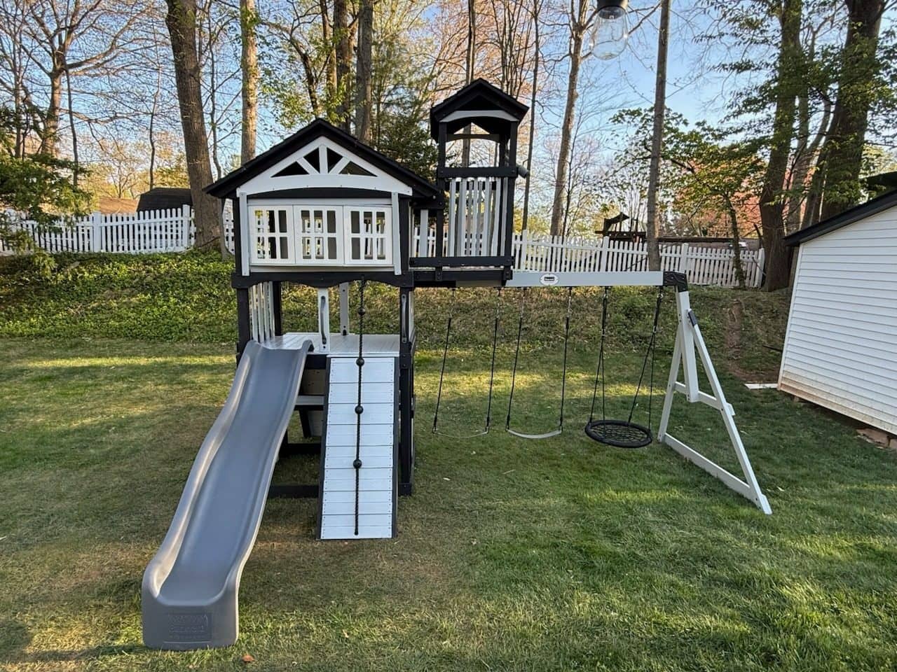 White and black wooden playset with a slide, climbing wall and swings in a fenced yard