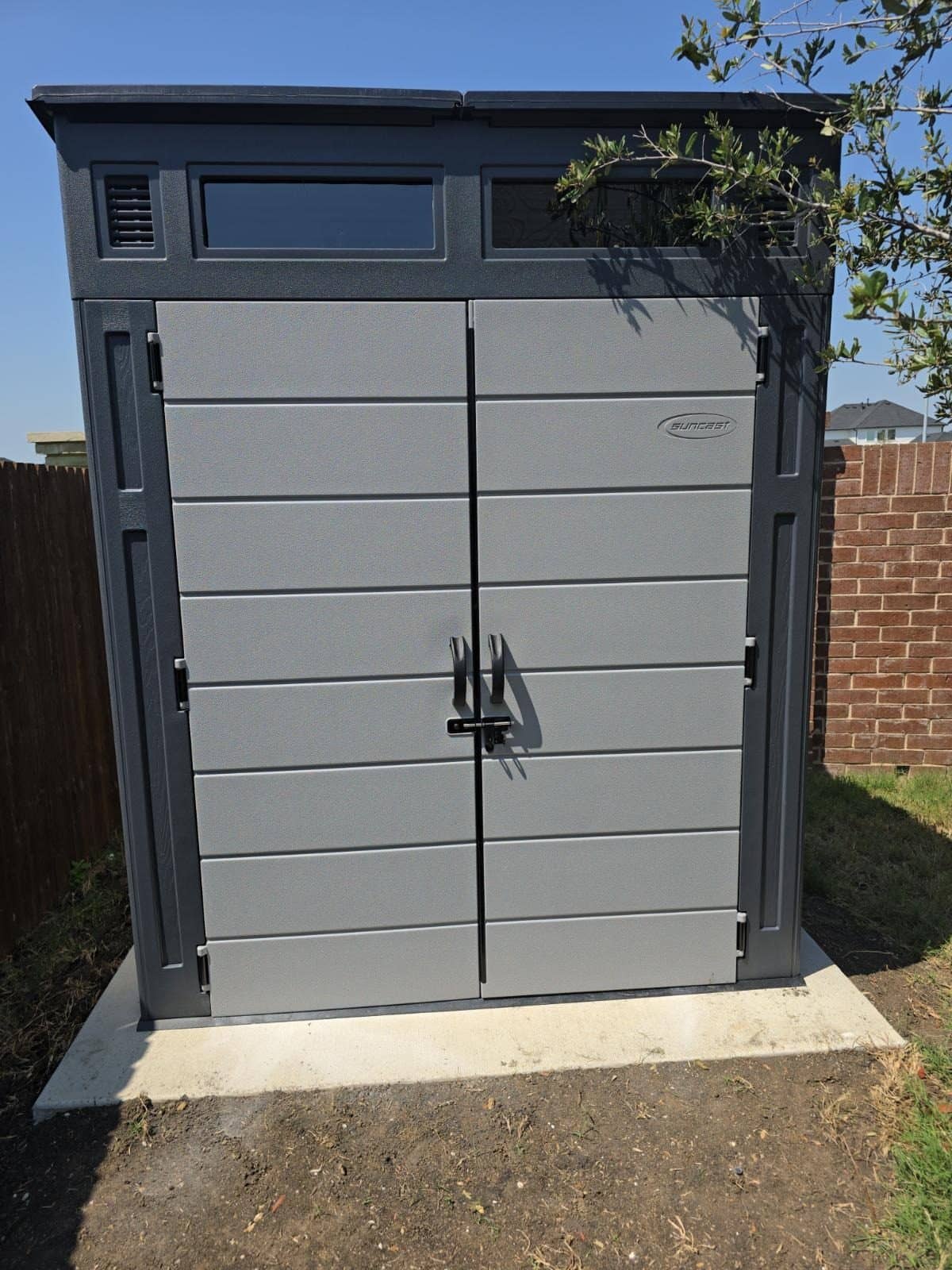 Gray resin storage shed with double doors standing on a concrete pad beside a brick wall