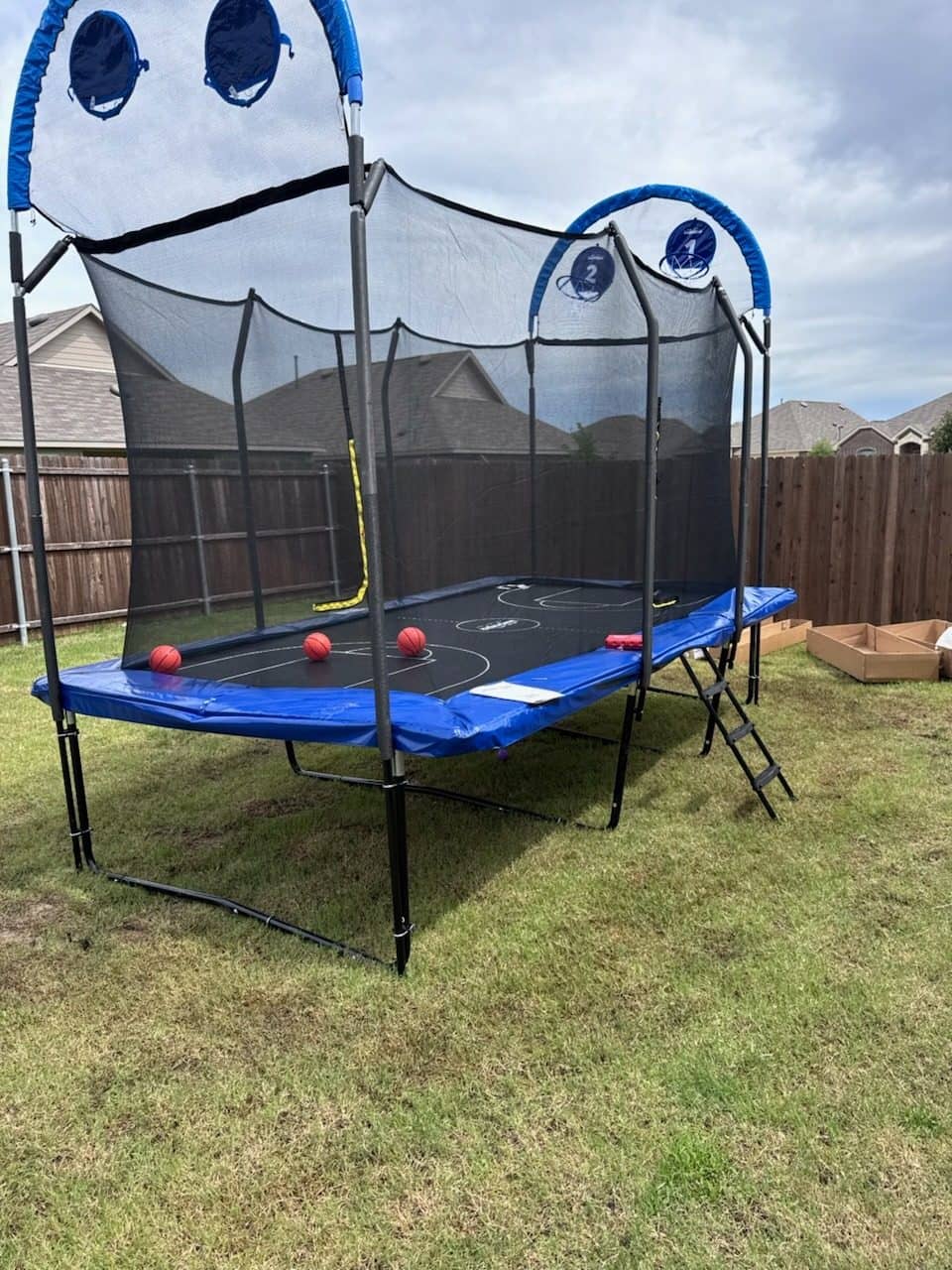 Rectangular trampoline with a blue pad, safety net and basketball hoop on a backyard lawn