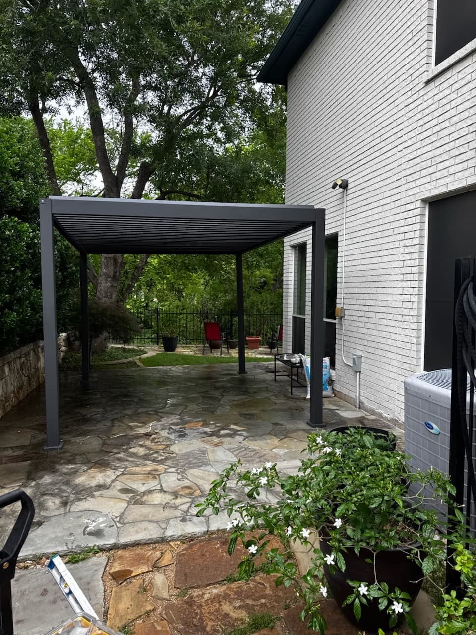 Dark louvered aluminium pergola over a flagstone patio beside a white brick house